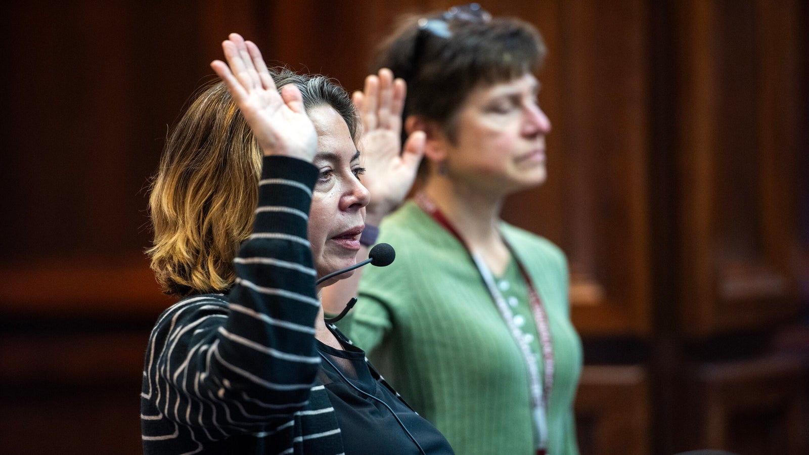 Two women stand with raised hands, one speaking into a headset microphone, appearing to participate in a formal proceeding or ceremony indoors.