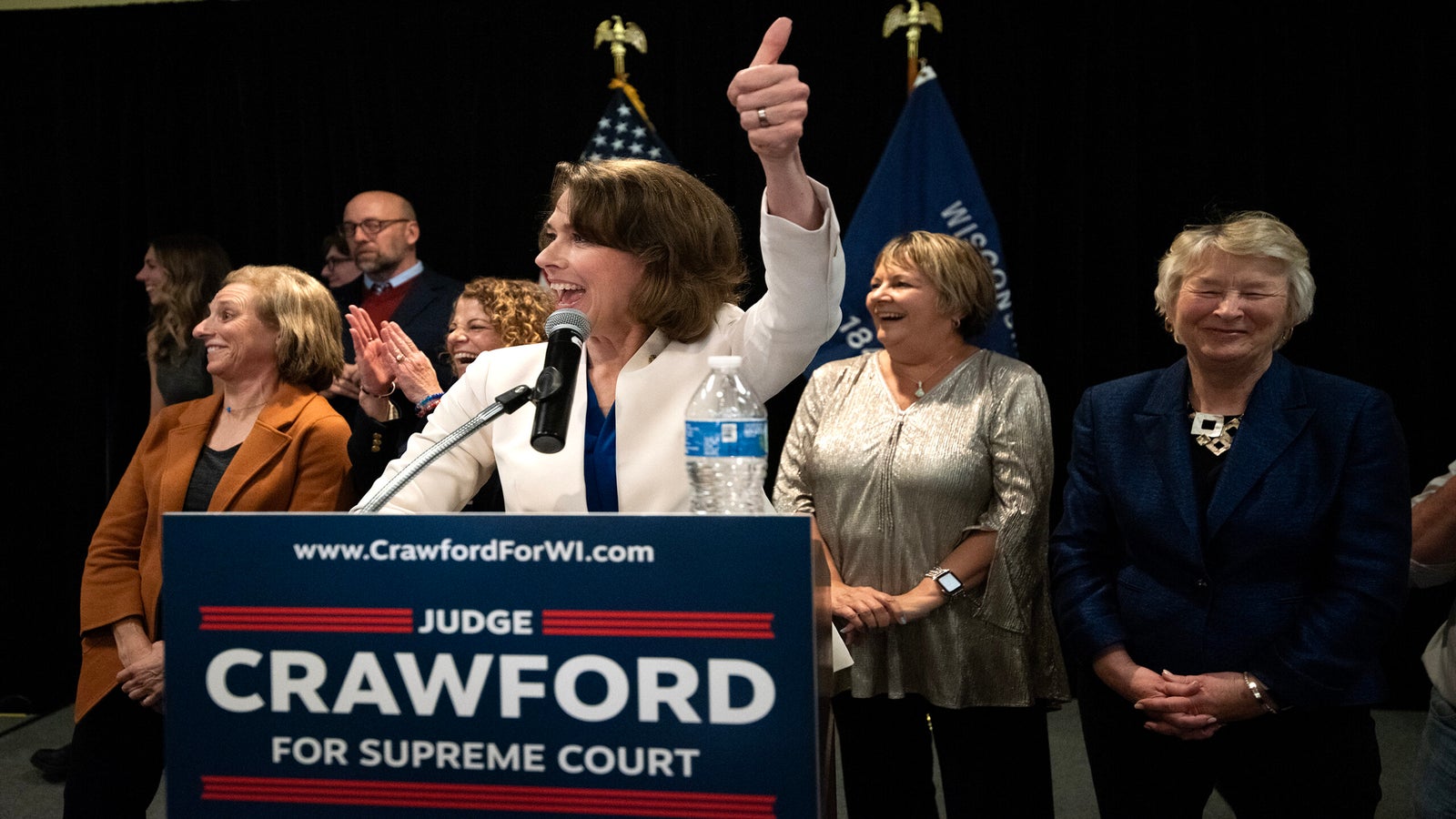 A woman stands at a podium with a Judge Crawford for Supreme Court sign, giving a thumbs up. Several people stand behind her, smiling, with flags displayed in the background.