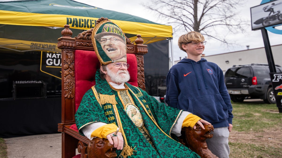 An older man in ornate green robes and a large hat with a face on it sits on a throne next to a boy standing, outdoors near a Packers tent and vehicles.