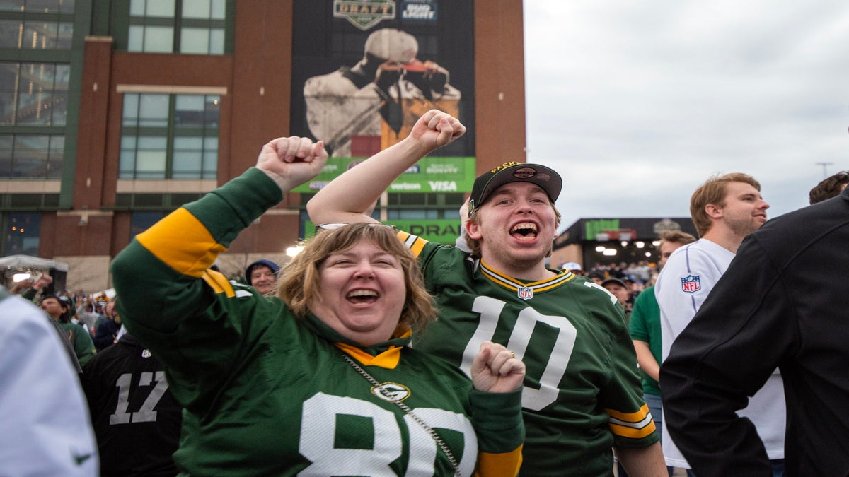 Two Green Bay Packers fans in team jerseys cheer among a crowd outside a stadium, with a large mural of a football player in the background.