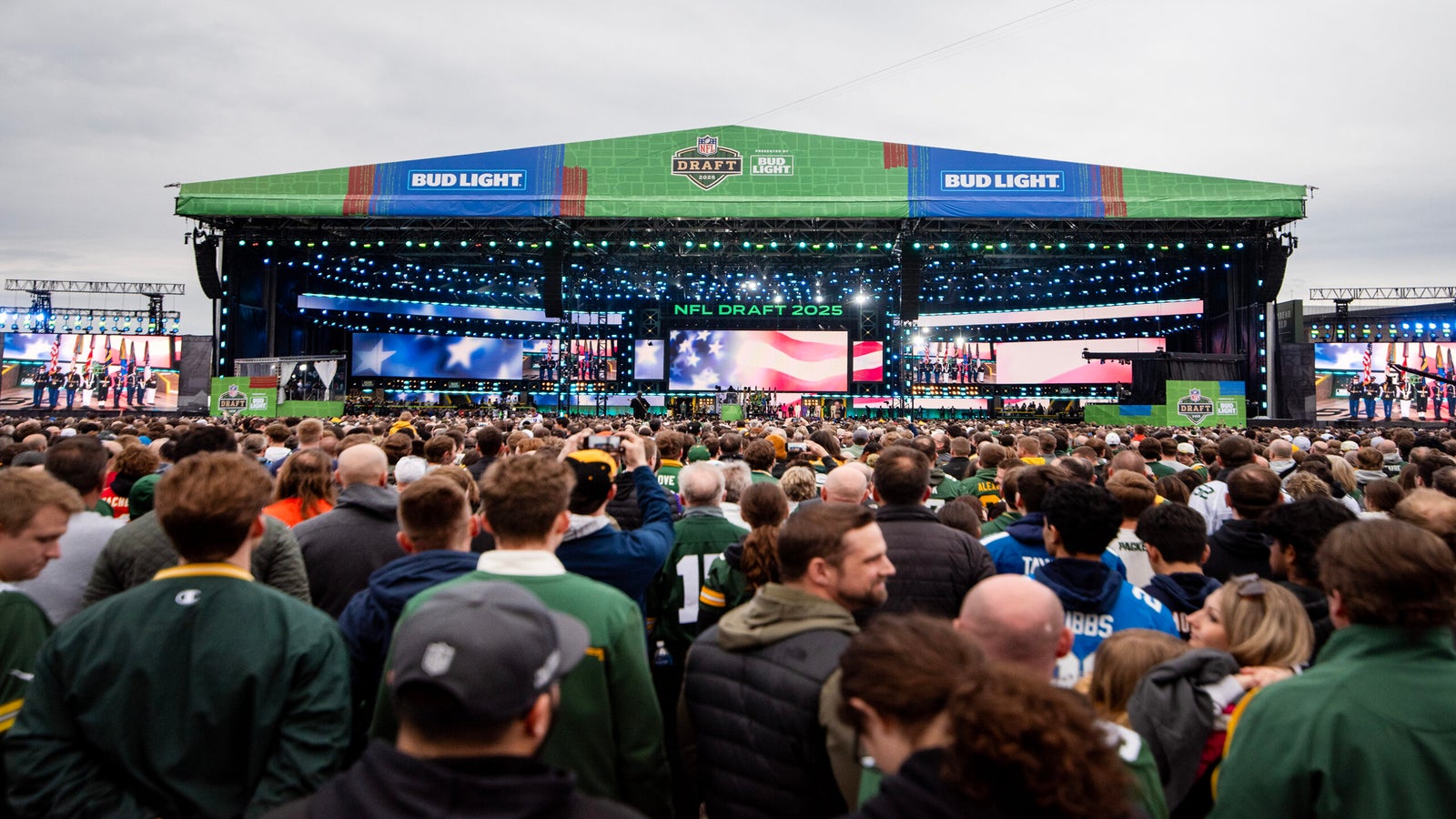 A large crowd gathers in front of the main stage at the NFL Draft 2025 event, with American flags displayed on the screens and stadium lights illuminating the scene.