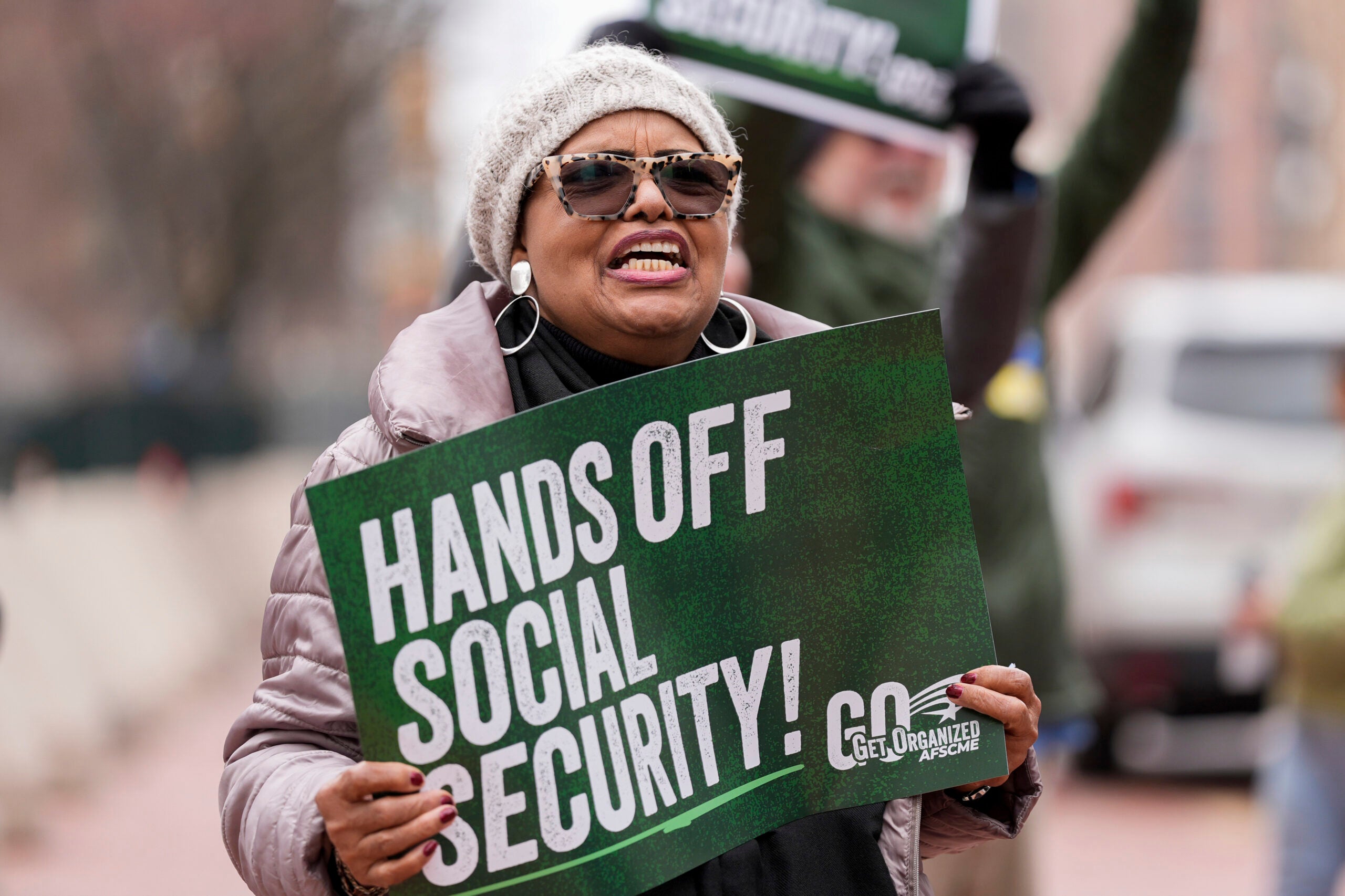 A person wearing sunglasses and a winter hat holds a Hands off Social Security! sign, participating in a protest.