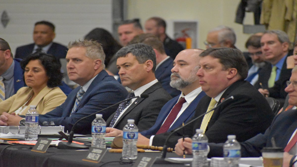 People in formal attire sitting at a conference table with nameplates and water bottles, attentively listening.