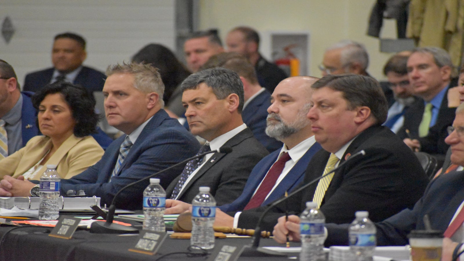 People in formal attire sitting at a conference table with nameplates and water bottles, attentively listening.