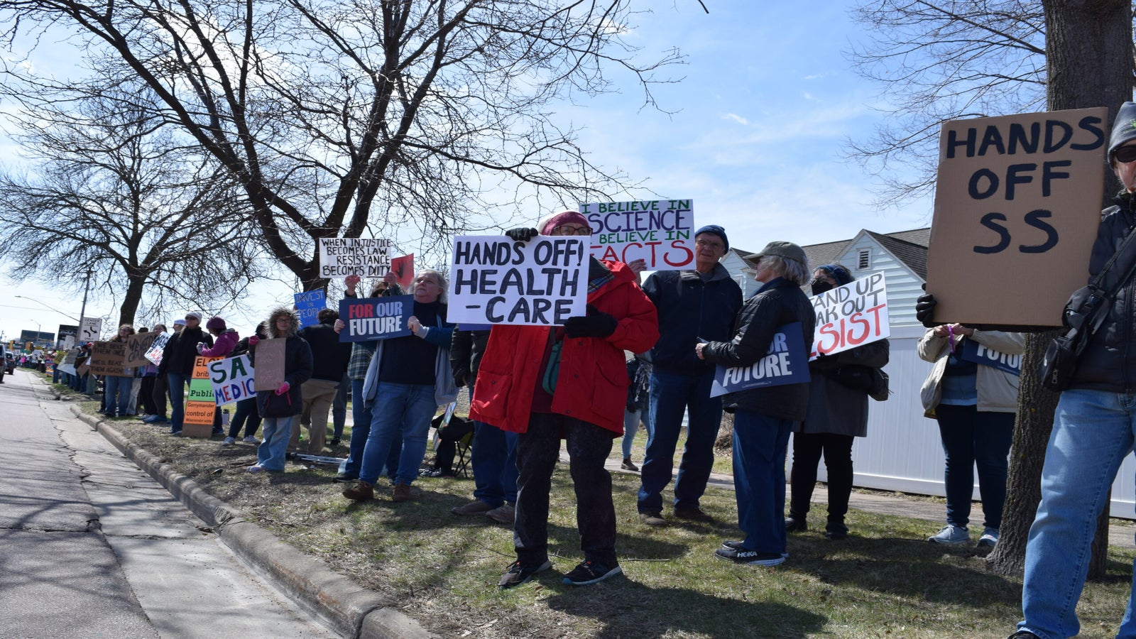 A group of people are gathered outdoors holding various protest signs, including messages about healthcare, science, and other social issues.