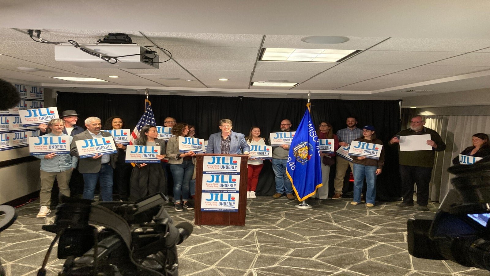 A speaker at a podium surrounded by people holding signs that read Jill for Justice, with an American and Wisconsin state flag in the background.