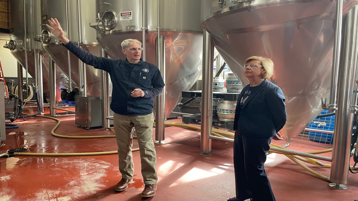 A man gestures while speaking to a woman in a brewery, surrounded by large metal fermentation tanks.