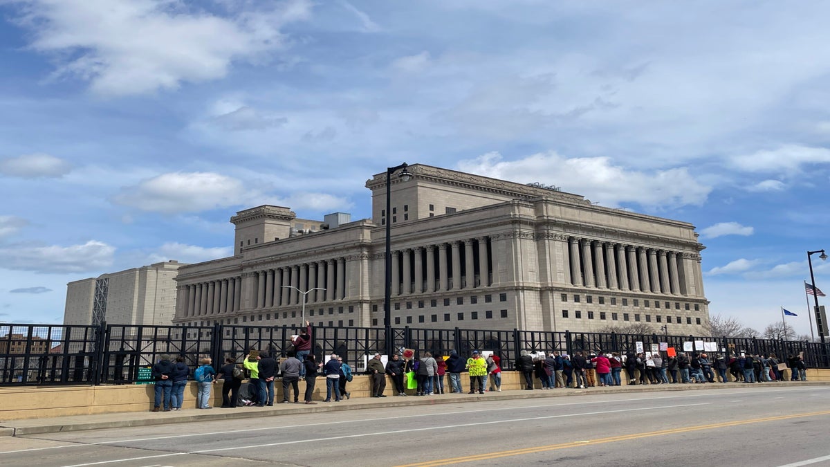 A group of people stand with signs in front of a large stone government building on a cloudy day.