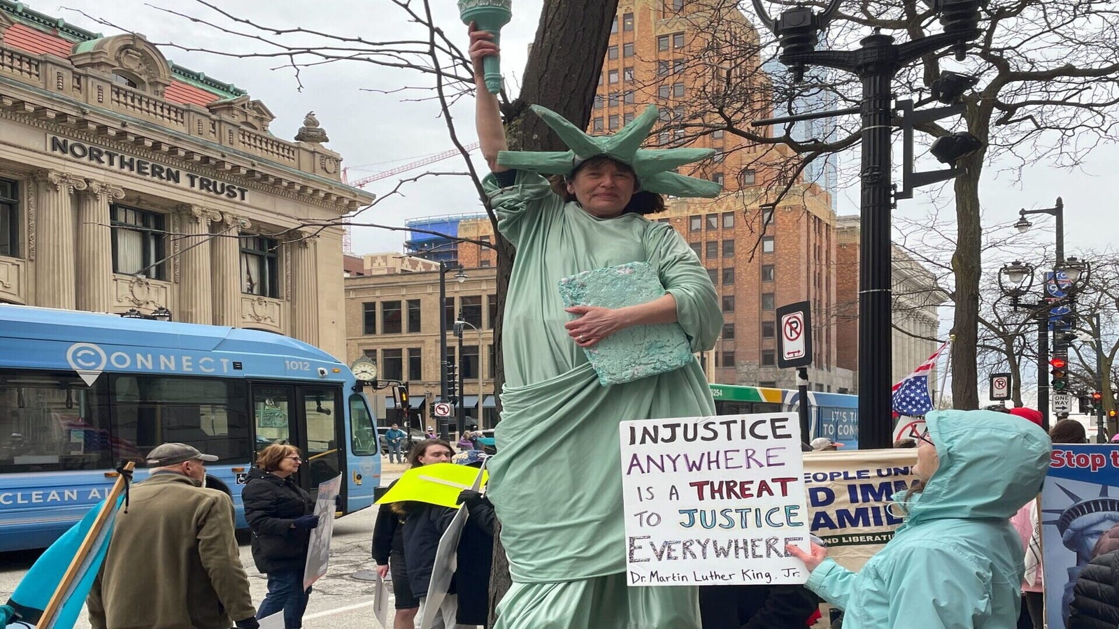 Person dressed as the Statue of Liberty stands on stilts holding a sign with a Martin Luther King Jr. quote at a public protest in an urban area. Other people hold signs nearby.