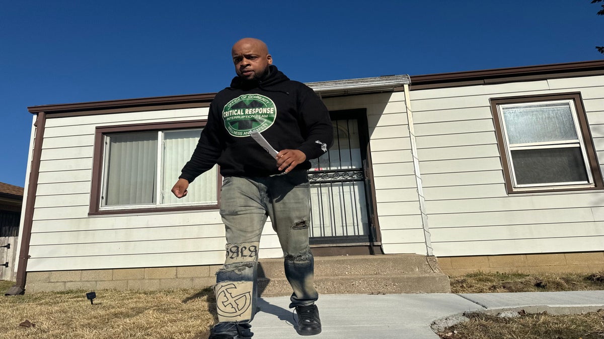 Man in a black hoodie and jeans walking on a path in front of a single-story house with white siding and a metal door.