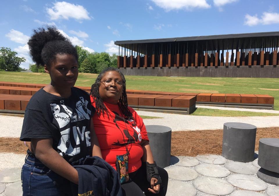 Two people sit and stand together outside the National Memorial for Peace and Justice in Montgomery, Alabama, with memorial columns visible in the background.