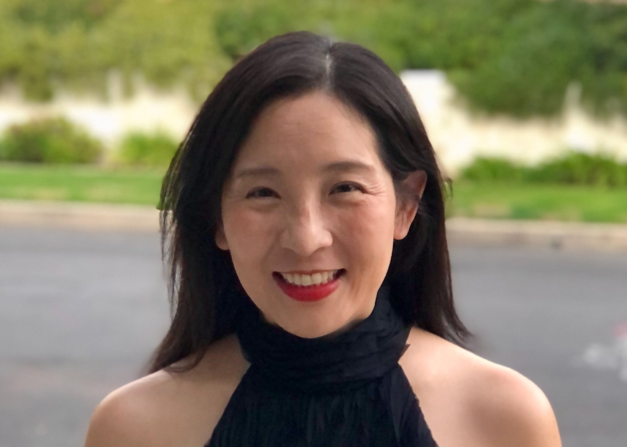 A woman with long dark hair, wearing a sleeveless black top, smiles while standing outdoors with greenery in the background.