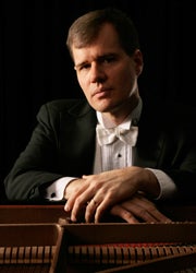 A man in a black tuxedo with a white bow tie sits at a grand piano, resting his arms on the instrument, against a dark background.