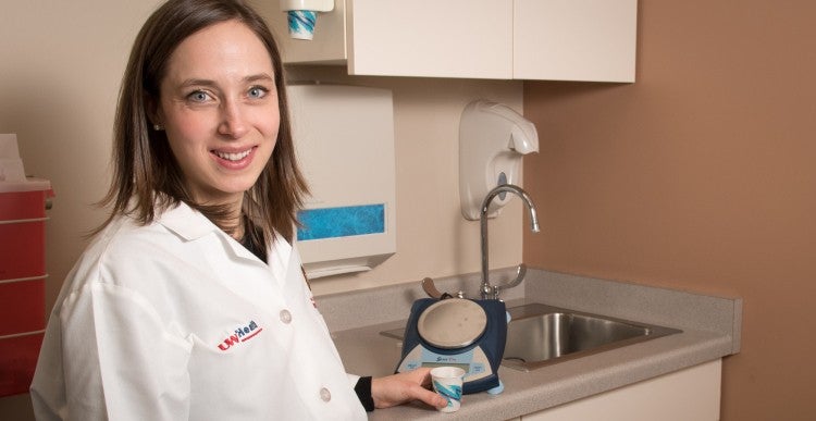 A person in a white coat stands in a medical or laboratory setting next to a small industrial device, holding a cup, and smiling at the camera.