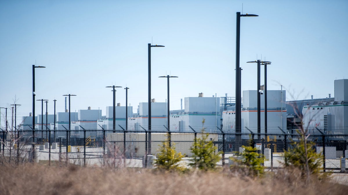 A row of white industrial buildings behind a chain-link fence with streetlights and sparse vegetation in the foreground.