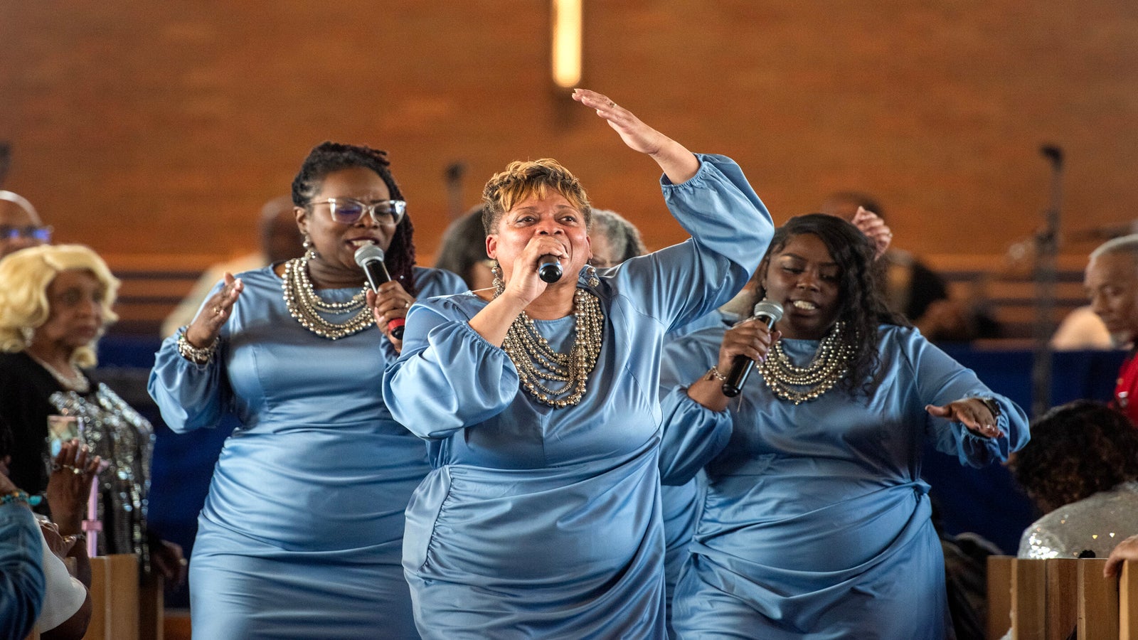 Three women in matching blue dresses sing passionately on stage in front of a church congregation, with a cross visible in the background.