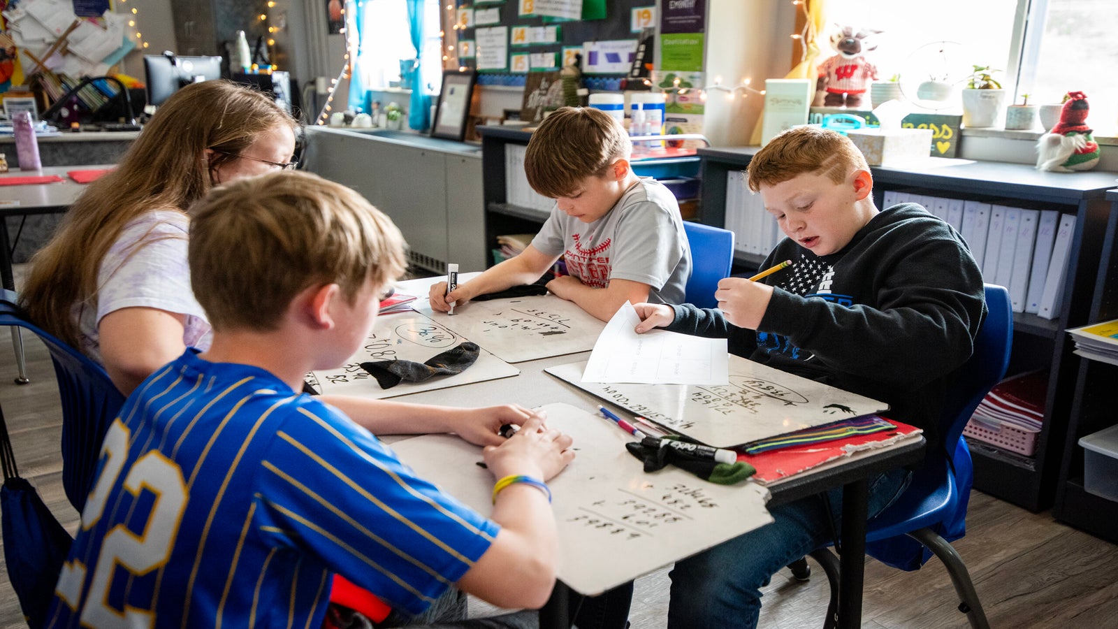 Four students sit at a table in a classroom, working on math problems using whiteboards and markers.