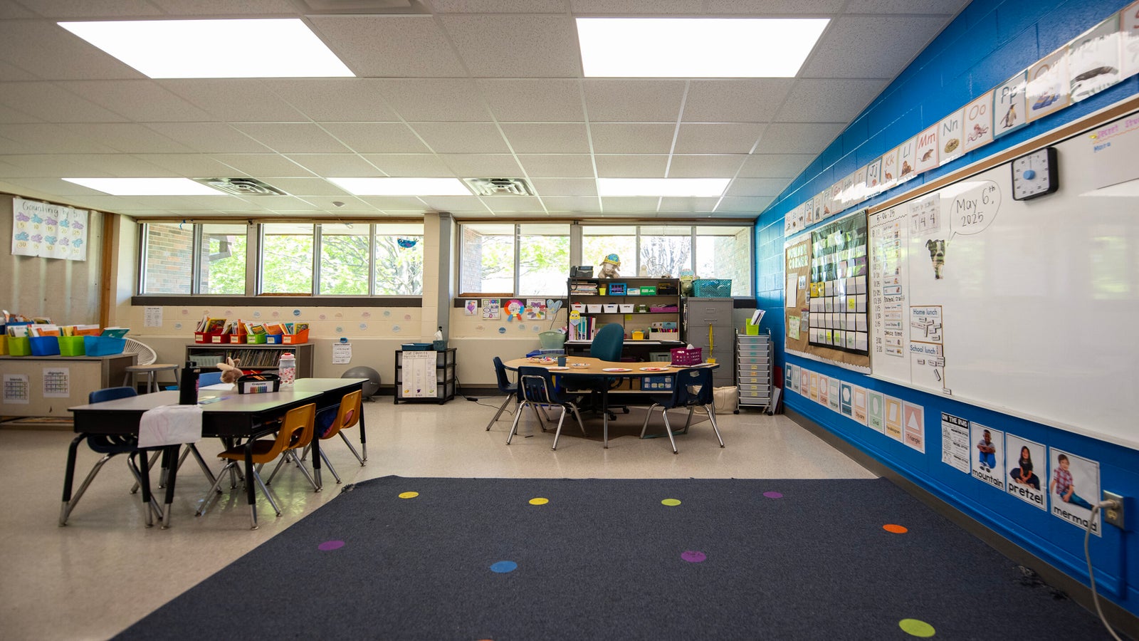 A brightly lit elementary classroom with desks, chairs, shelves of supplies, a large whiteboard, and colorful decorations on the walls.