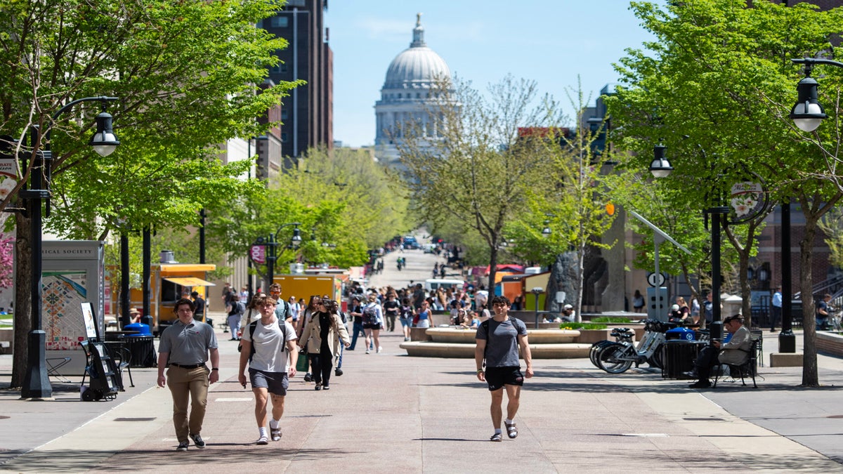 People walk along a tree-lined pedestrian street with the Wisconsin State Capitol building visible in the background on a sunny day.