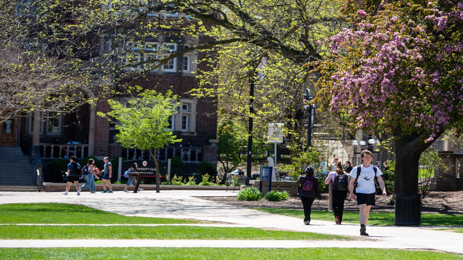 Students walk along a paved path lined with grass and blooming trees on a college campus, with a brick building visible in the background.