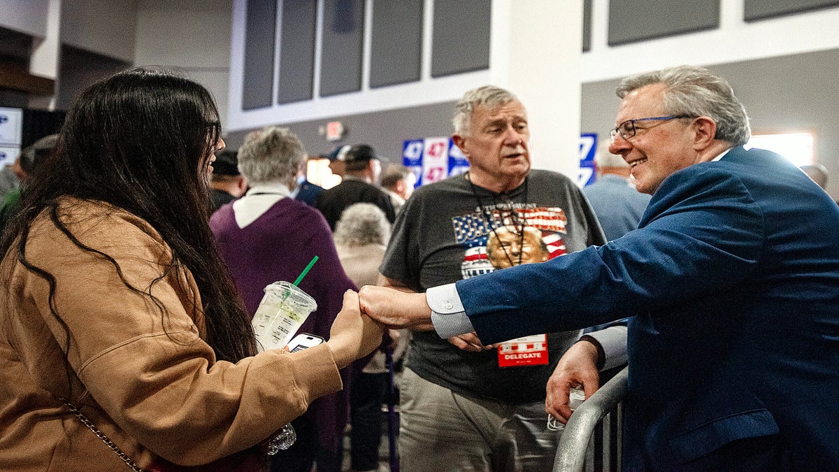 A man in a blue suit shakes hands with a woman holding a drink while another man stands nearby at a crowded indoor event.