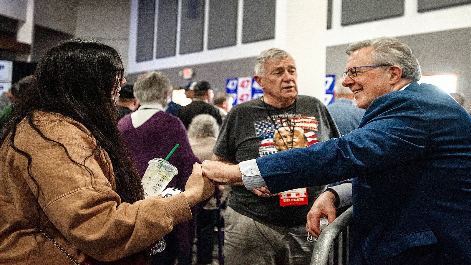 A man in a blue suit shakes hands with a woman holding a drink while another man stands nearby at a crowded indoor event.