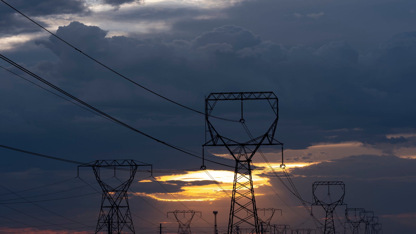 High-voltage power lines and transmission towers are silhouetted against a cloudy sky at sunset.