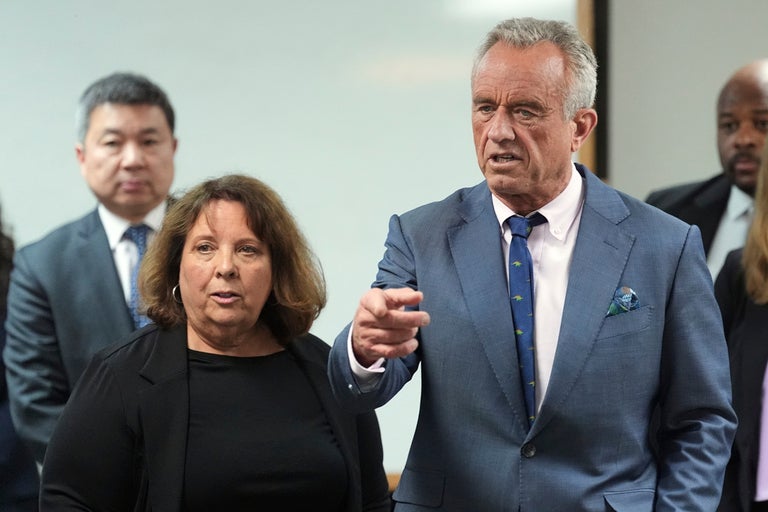 A man in a blue suit gestures while speaking, standing next to a woman in black, with three other people in business attire in the background.