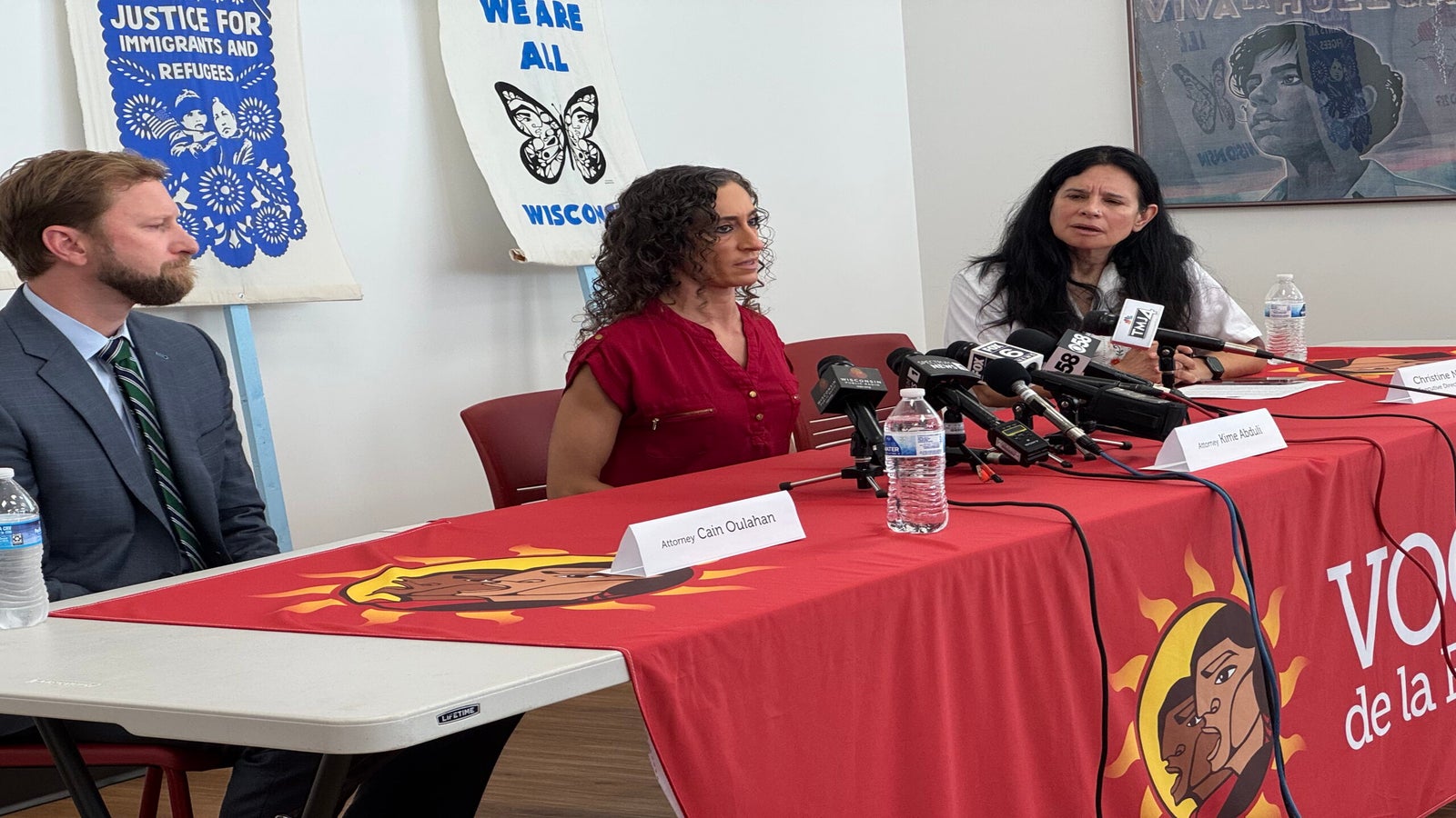 Three people sit at a table with microphones during a press conference, in front of banners supporting immigrants and refugees.