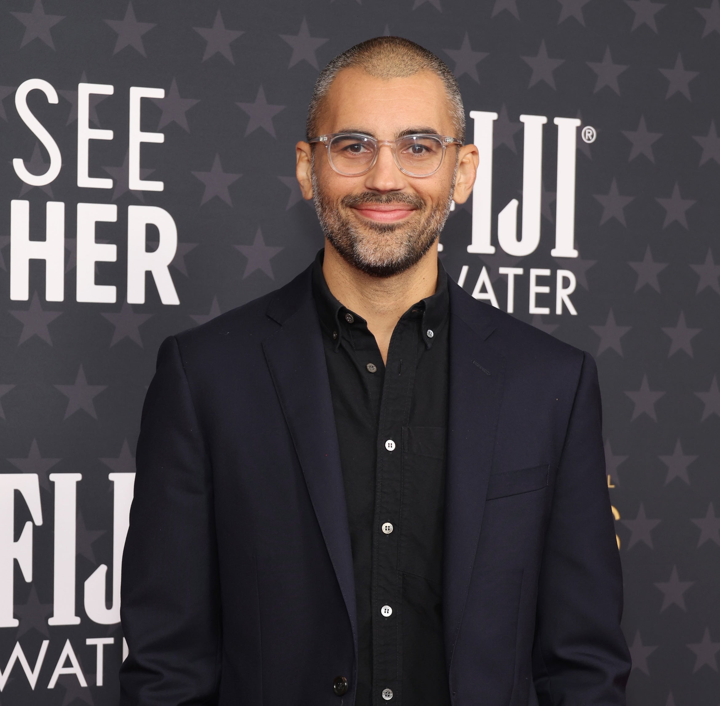 A man in a black suit and glasses stands in front of a step-and-repeat backdrop featuring the Fiji Water logo and star patterns.