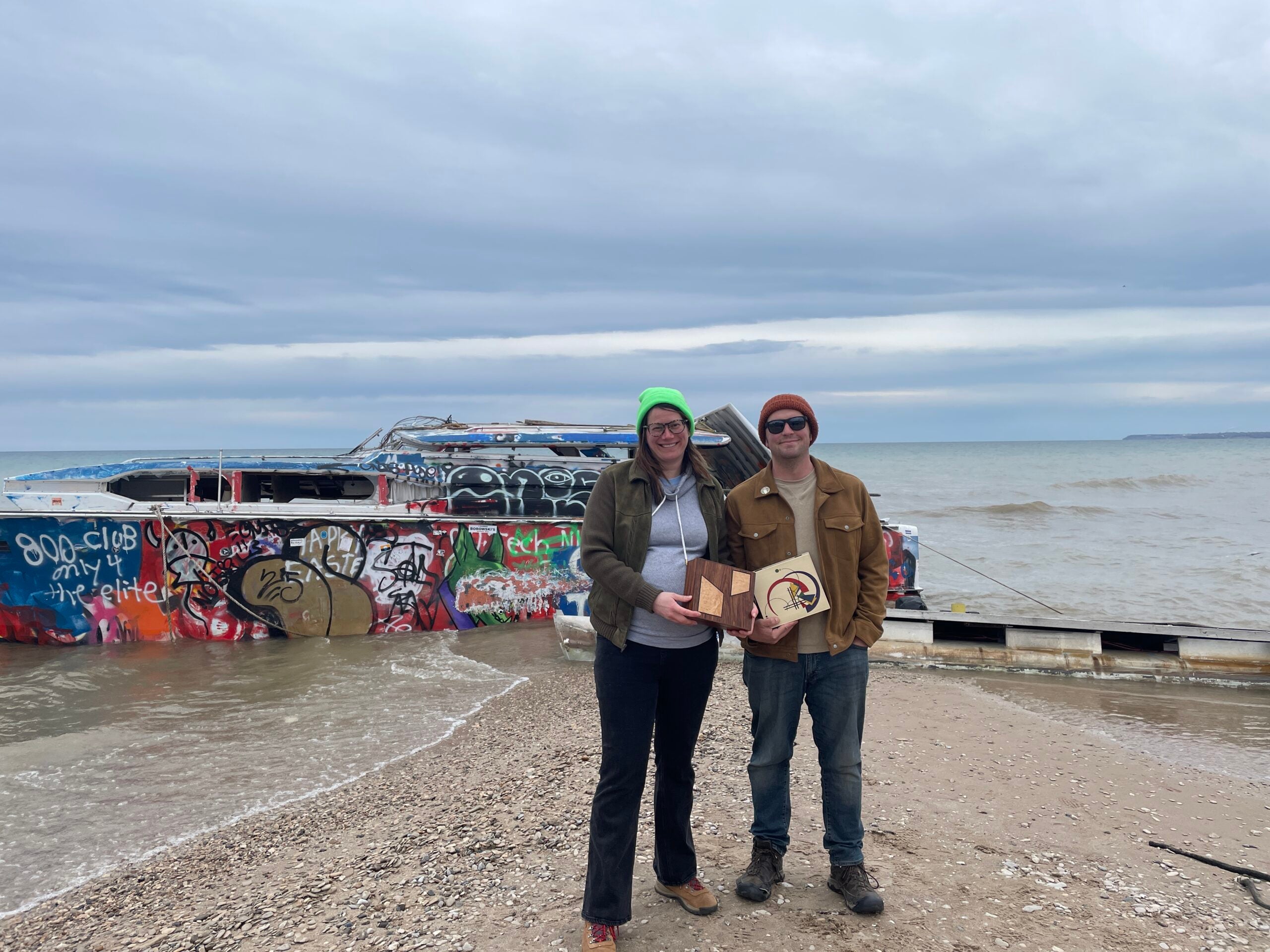 Two people stand on a rocky beach in front of a large, graffiti-covered, partially sunken boat under a cloudy sky.