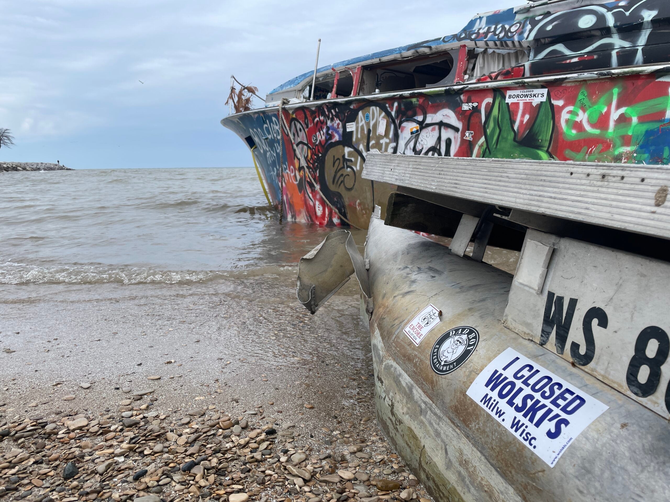 A graffiti-covered, abandoned boat sits on a rocky shore with a metal pipe in the foreground displaying various stickers.