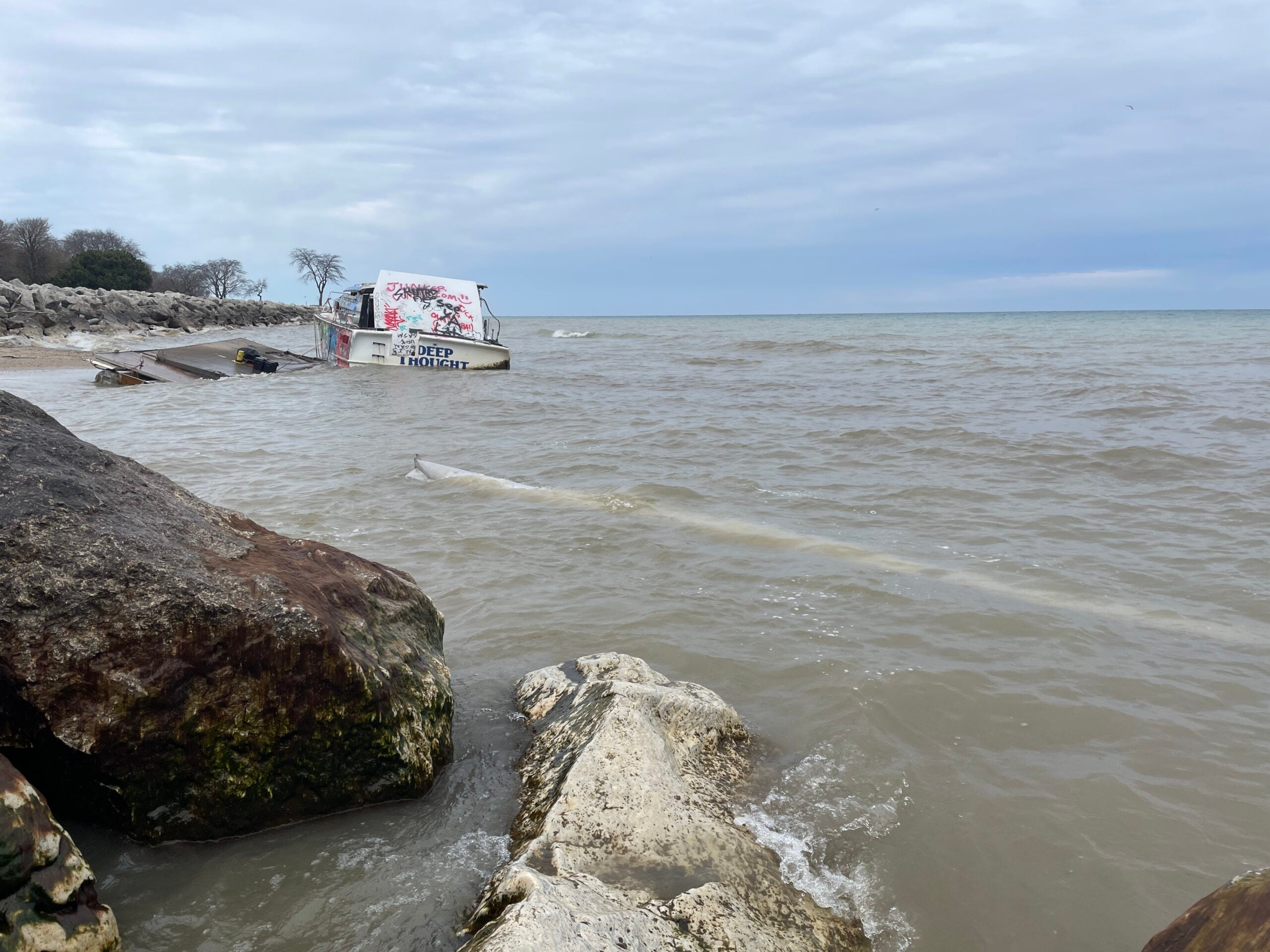 A partially submerged, graffiti-covered structure sits near the rocky shoreline of a large body of water under a cloudy sky.