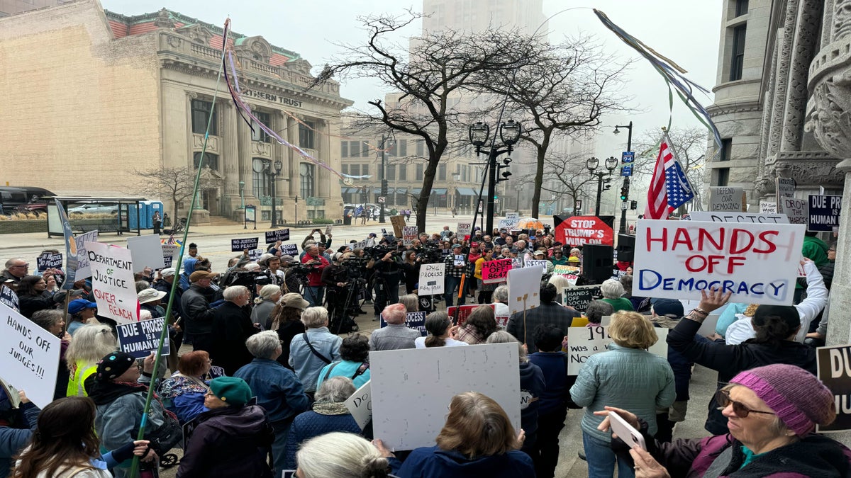 A crowd of protesters gathers on a city street holding signs with messages about democracy, justice, and human rights. Reporters with cameras are present in the center.