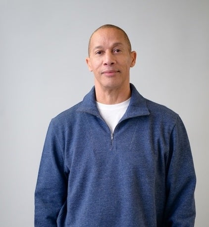 A man with short hair wearing a blue quarter-zip sweater over a white shirt stands against a plain light gray background.