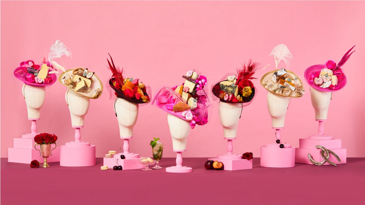 Seven mannequin heads display decorative hats adorned with desserts and feathers, set against a pink background with various props like roses, fruit, and champagne glasses.