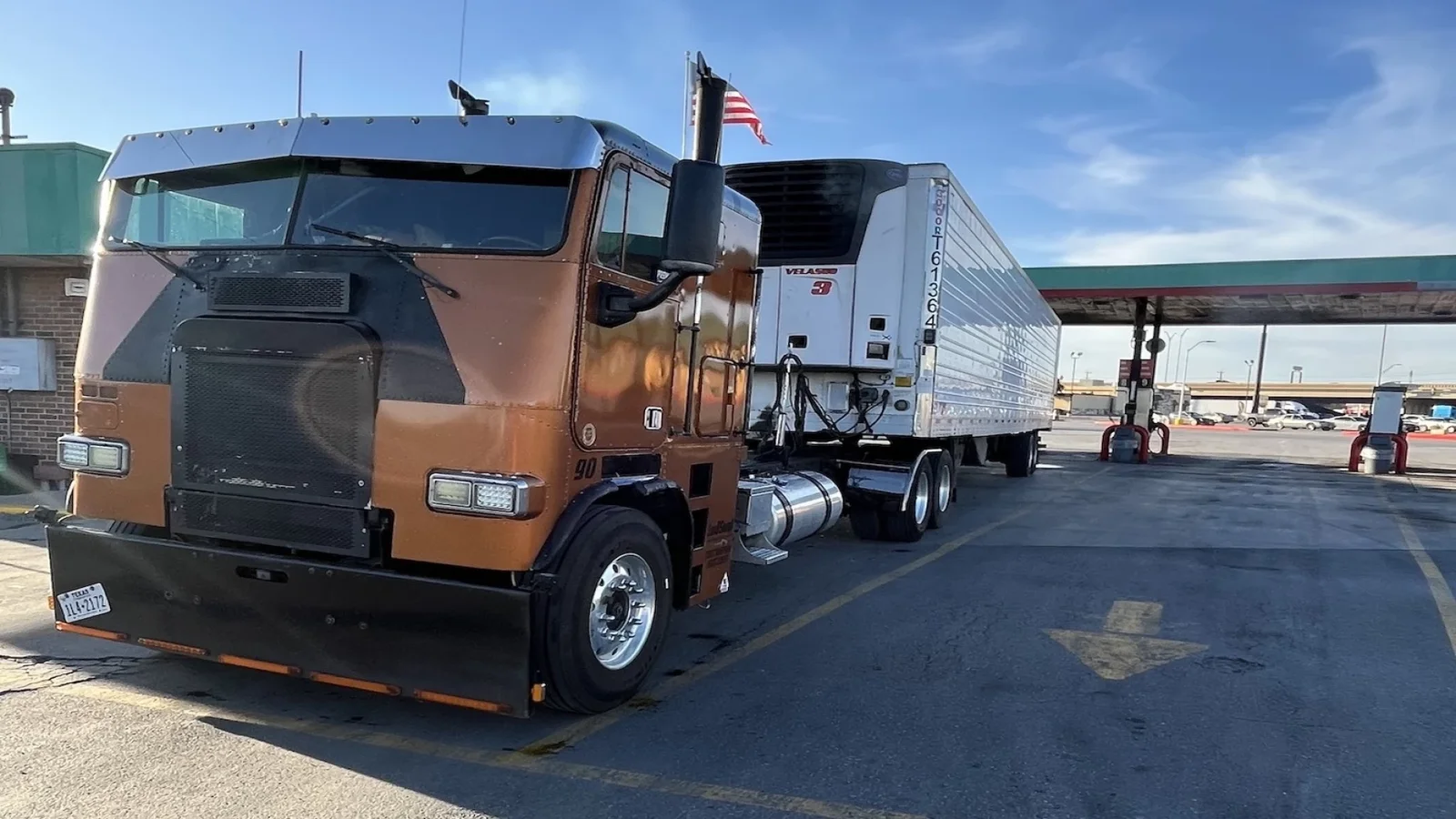A brown and black semi-truck with a trailer is parked at a fuel station under a clear sky. An American flag is attached to the truck.