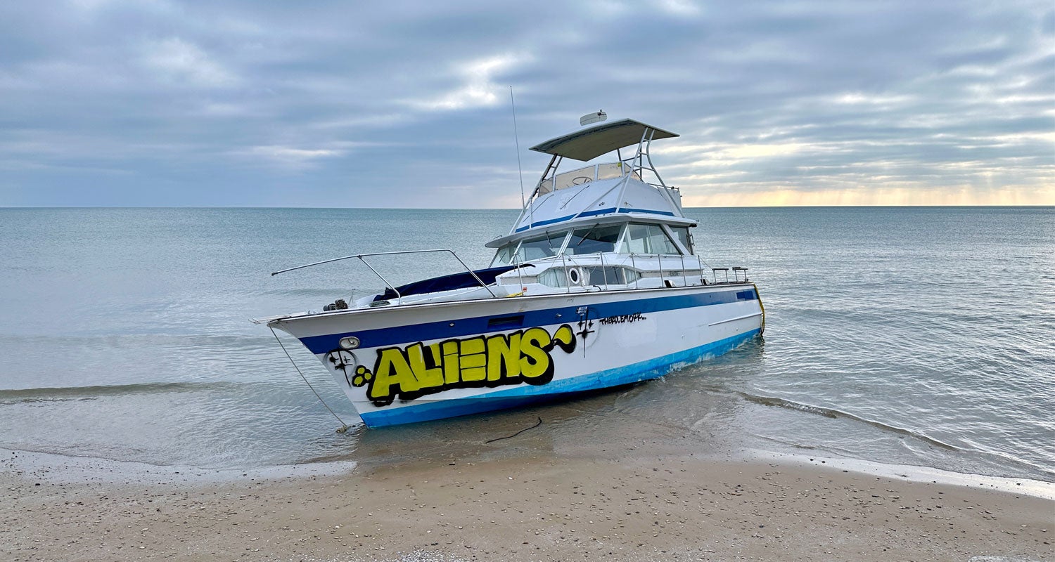 A white and blue boat with ALIENS spray-painted on the side is beached on a sandy shore under a cloudy sky.