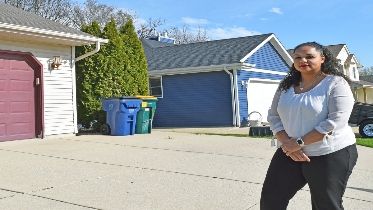 A woman stands on a driveway in front of a house with a maroon garage door, recycling and trash bins, and another blue house in the background.