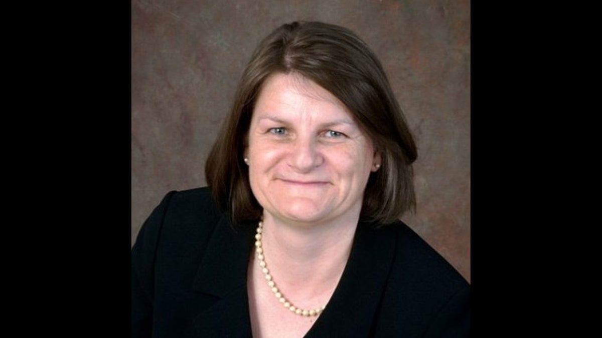A woman with straight brown hair wearing a black blazer and a pearl necklace, smiling against a brown mottled background.