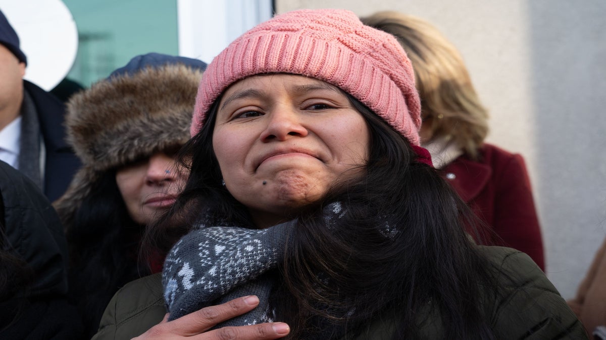 A woman wearing a pink knit hat and patterned scarf stands outdoors with a neutral yet emotional expression, surrounded by other people in winter clothing.