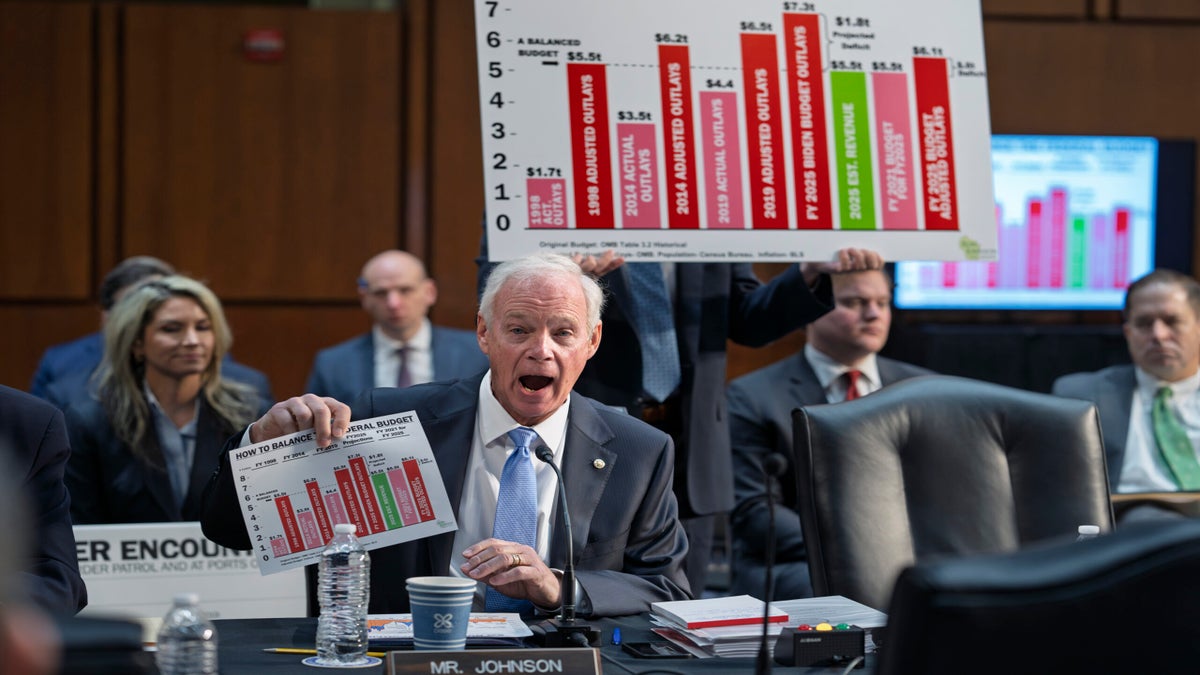 A man in a suit holds up a chart with budget data at a formal meeting, while another person in the background displays a larger version of the same chart.