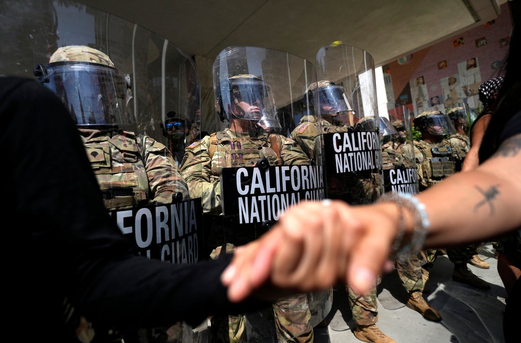 Several California National Guard members in riot gear and holding shields stand in a line as protesters in the foreground hold hands.