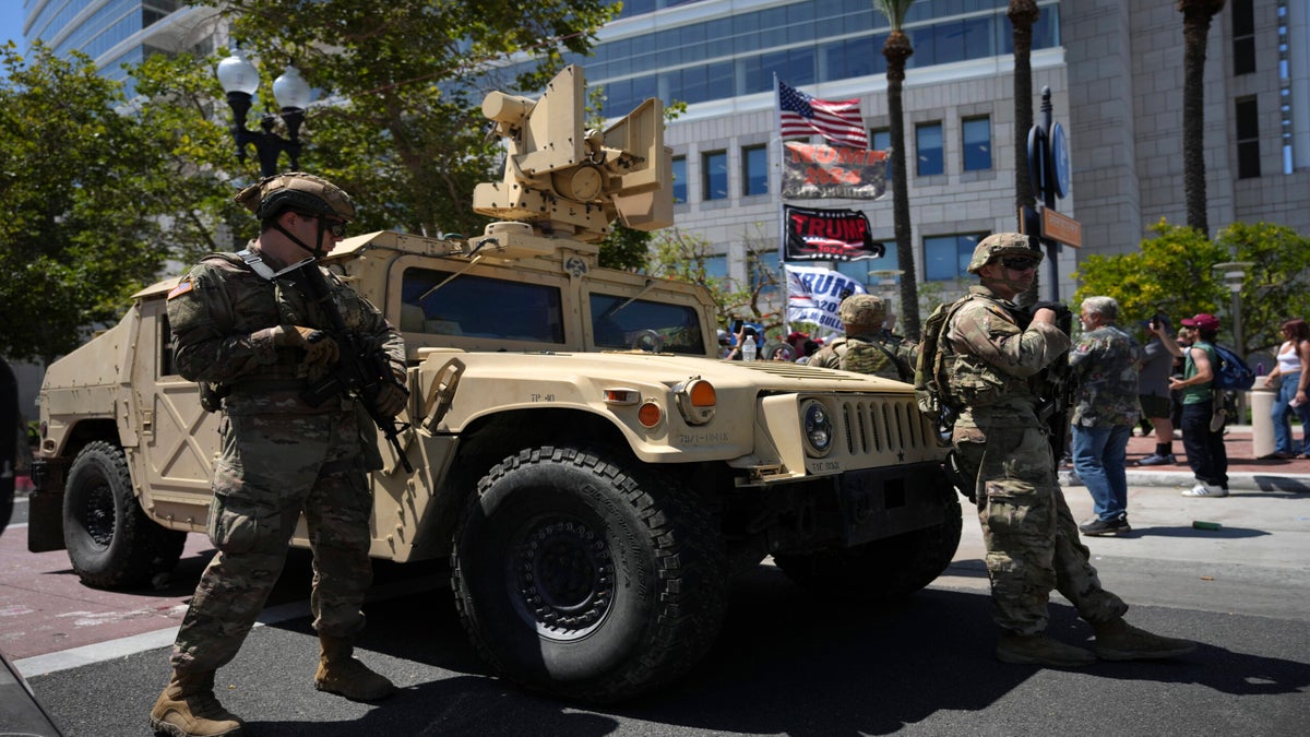 Armed soldiers stand beside an armored military vehicle on a city street as people gather nearby, with flags and banners visible in the background.