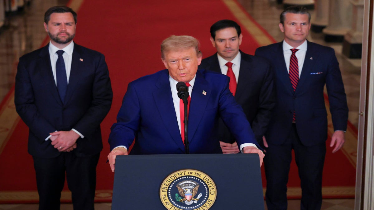 A man stands at a podium with the presidential seal, speaking into microphones, with three men in suits standing behind him in a formal hallway.