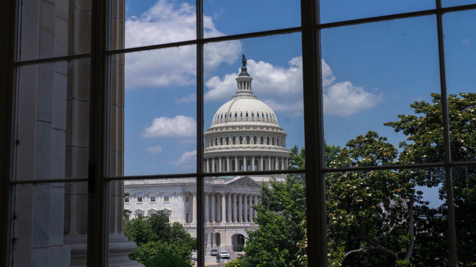 The U.S. Capitol building is seen through a window, framed by trees and a partly cloudy sky.