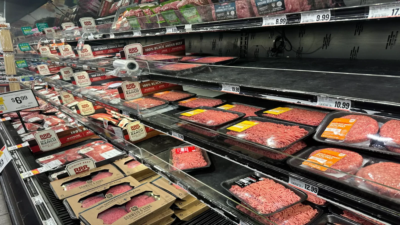 Packages of ground beef and other meat products are displayed on partially empty supermarket shelves, with price labels visible above each section.