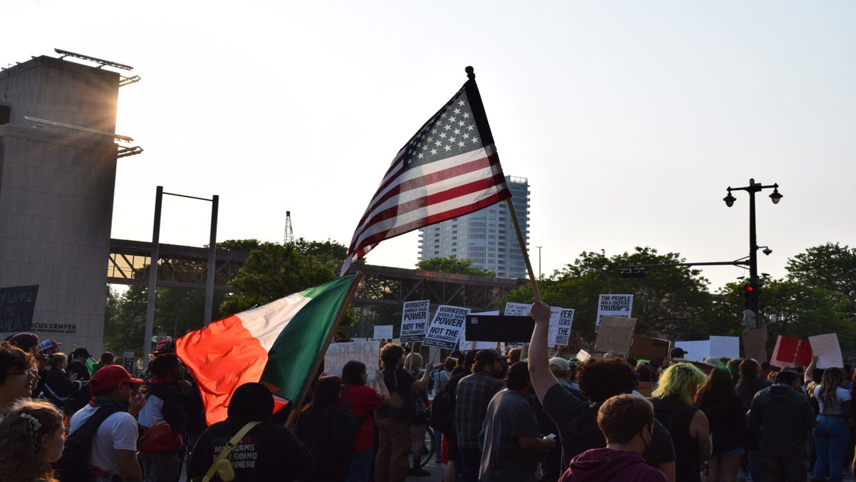 A crowd of protesters holds signs and waves American and Mexican flags during a daytime demonstration in an urban area.