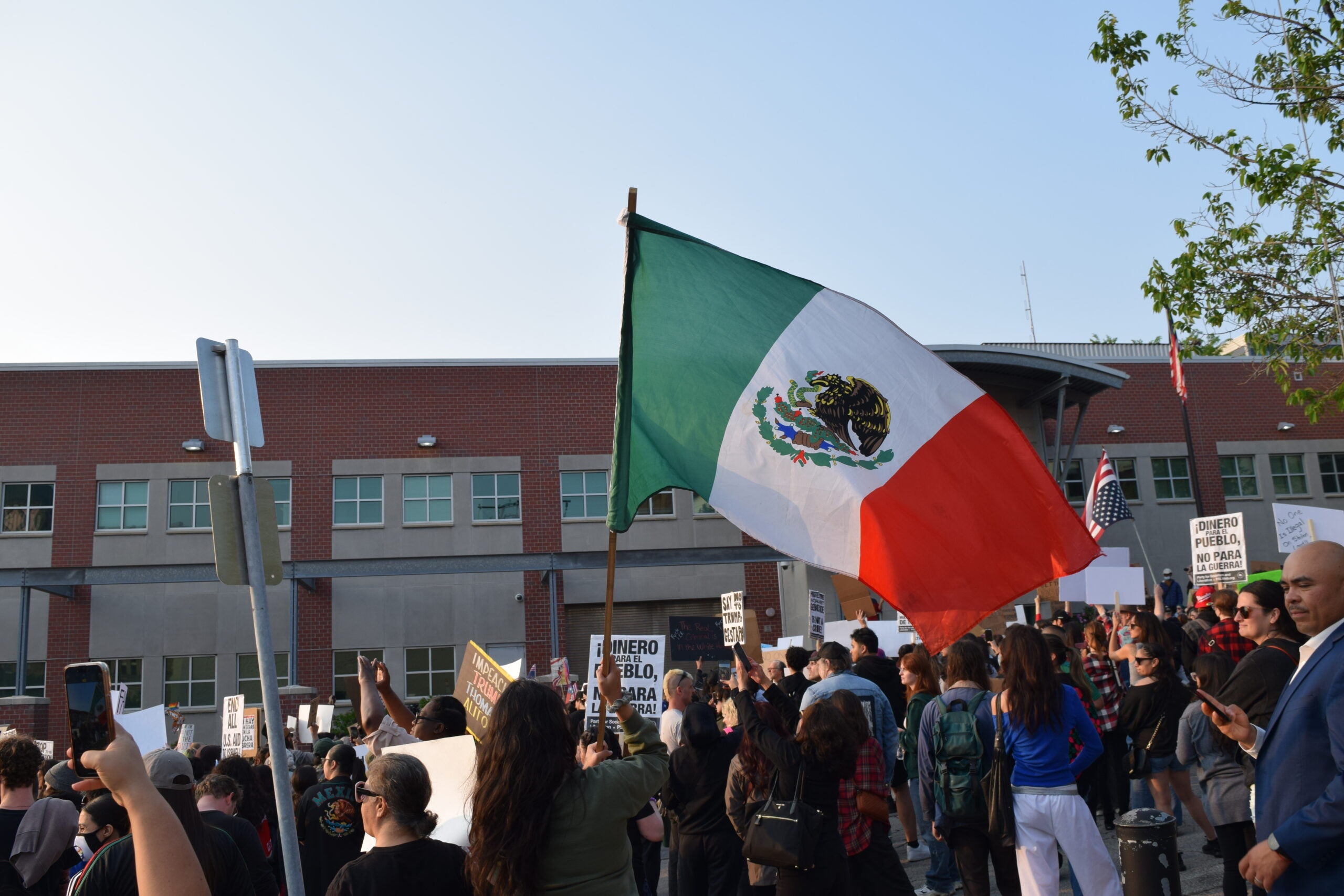 A crowd of people holding signs and a large Mexican flag gather in front of a brick building during a protest or demonstration.