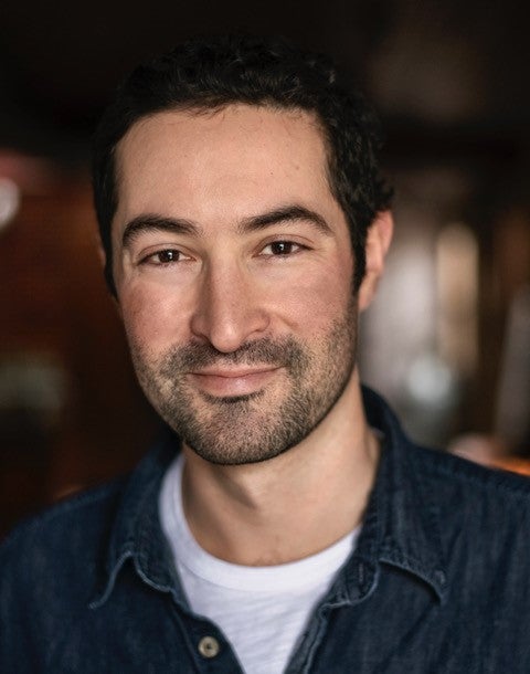 A man with short dark hair and a beard, wearing a dark denim shirt over a white t-shirt, stands indoors with a slight smile and a blurred background.